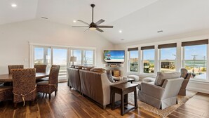 A spacious living room with a dining area, a ceiling fan, a TV, and large windows offering a view of the beach. - Endless Summer: 1-Min Walk Beach, Pool, Hot Tub (Kitty Hawk)