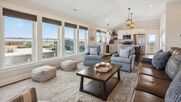 A spacious living room with a large window, a kitchen, and a view of the ocean. - Endless Summer: 1-Min Walk Beach, Pool, Hot Tub (Kitty Hawk)