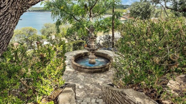 A stone fountain surrounded by greenery, with a view of a lake and buildings in the background.