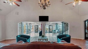 A spacious living room with a large brown leather sofa, two teal armchairs, a coffee table, and a chandelier. A flat-screen TV is mounted on the wall.