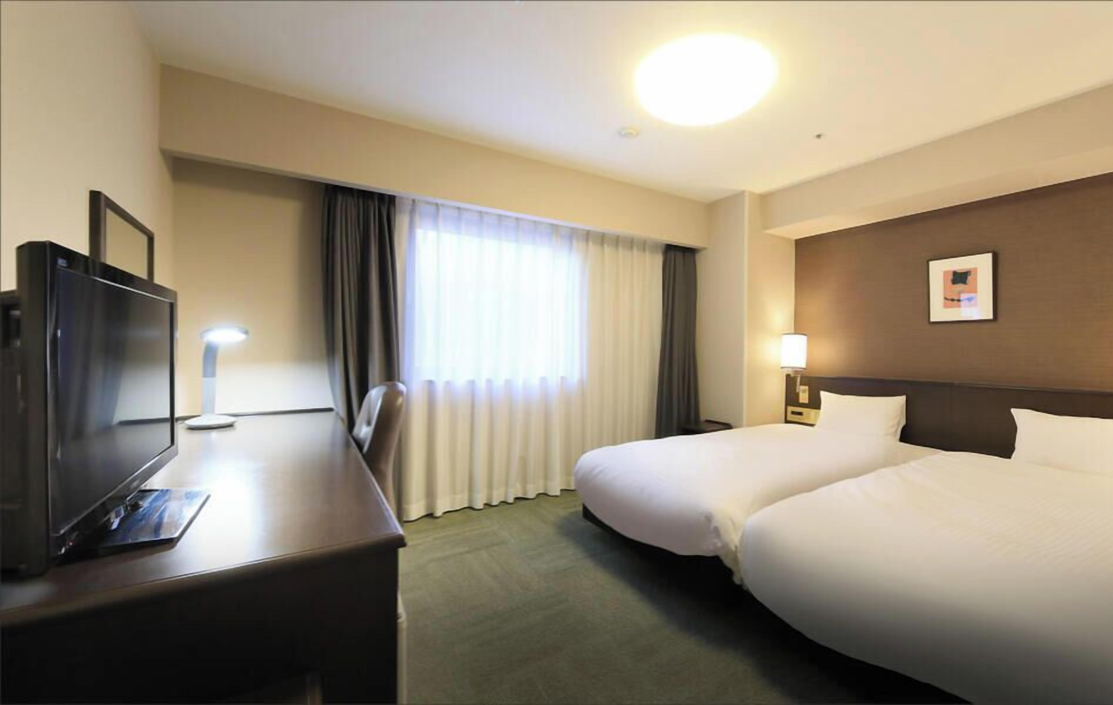 Quarto de hotel com cama, escrivaninha, televisão, abajur e quadro na parede.