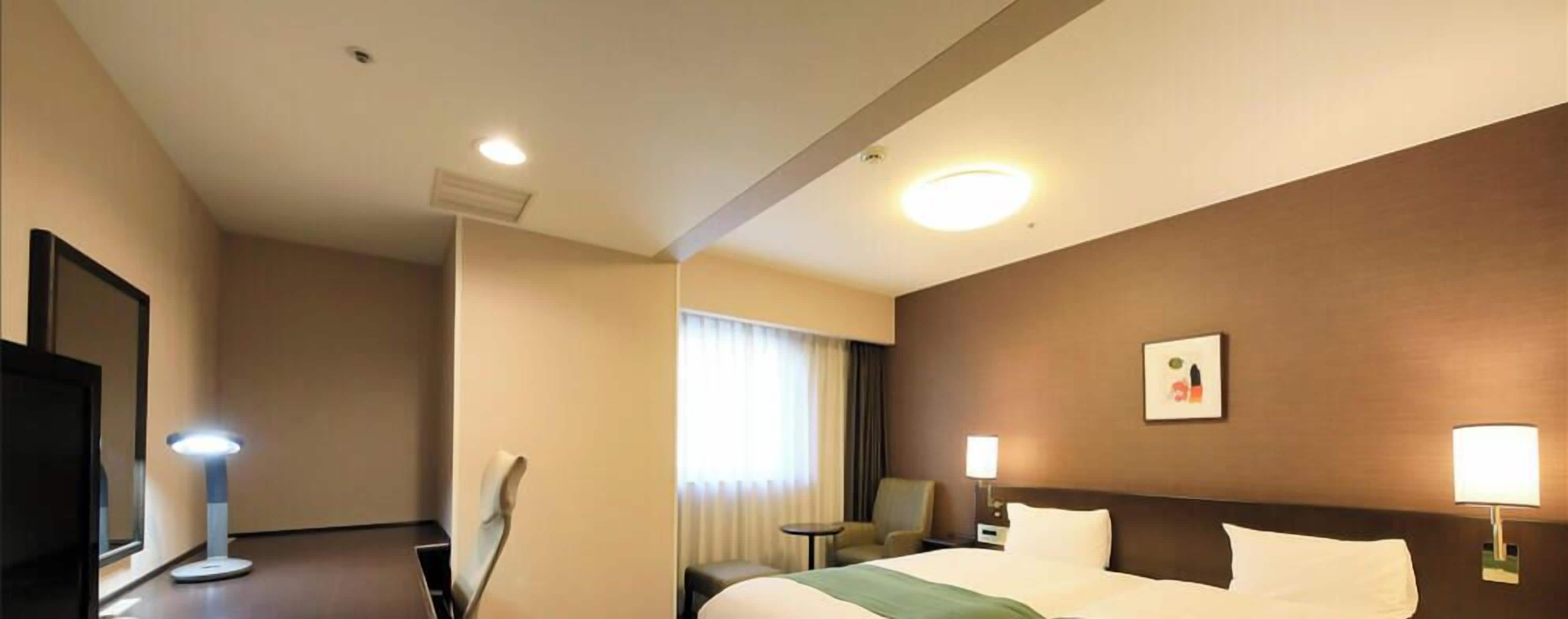 Quarto de hotel com cama, luminárias de cabeceira, uma poltrona, uma mesinha e um espelho.
