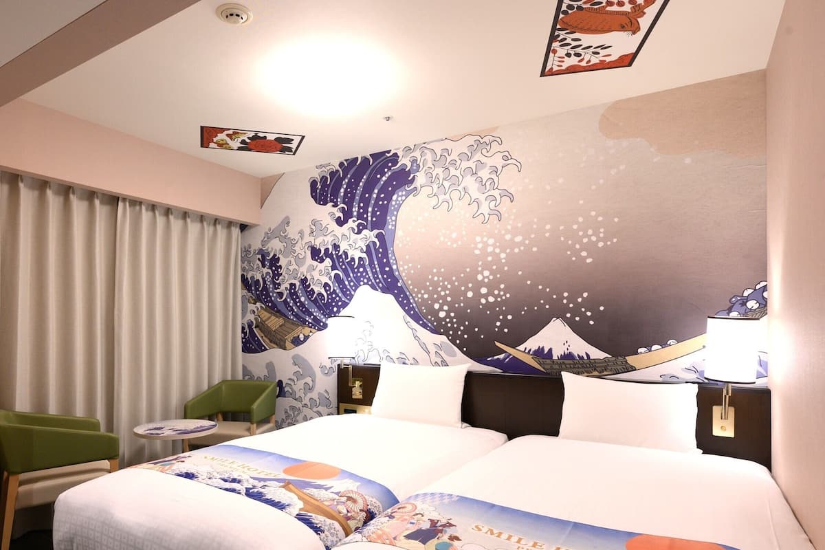 Une chambre d’hôtel avec un grand lit, un fauteuil vert, une petite table et une fresque murale représentant une vague.