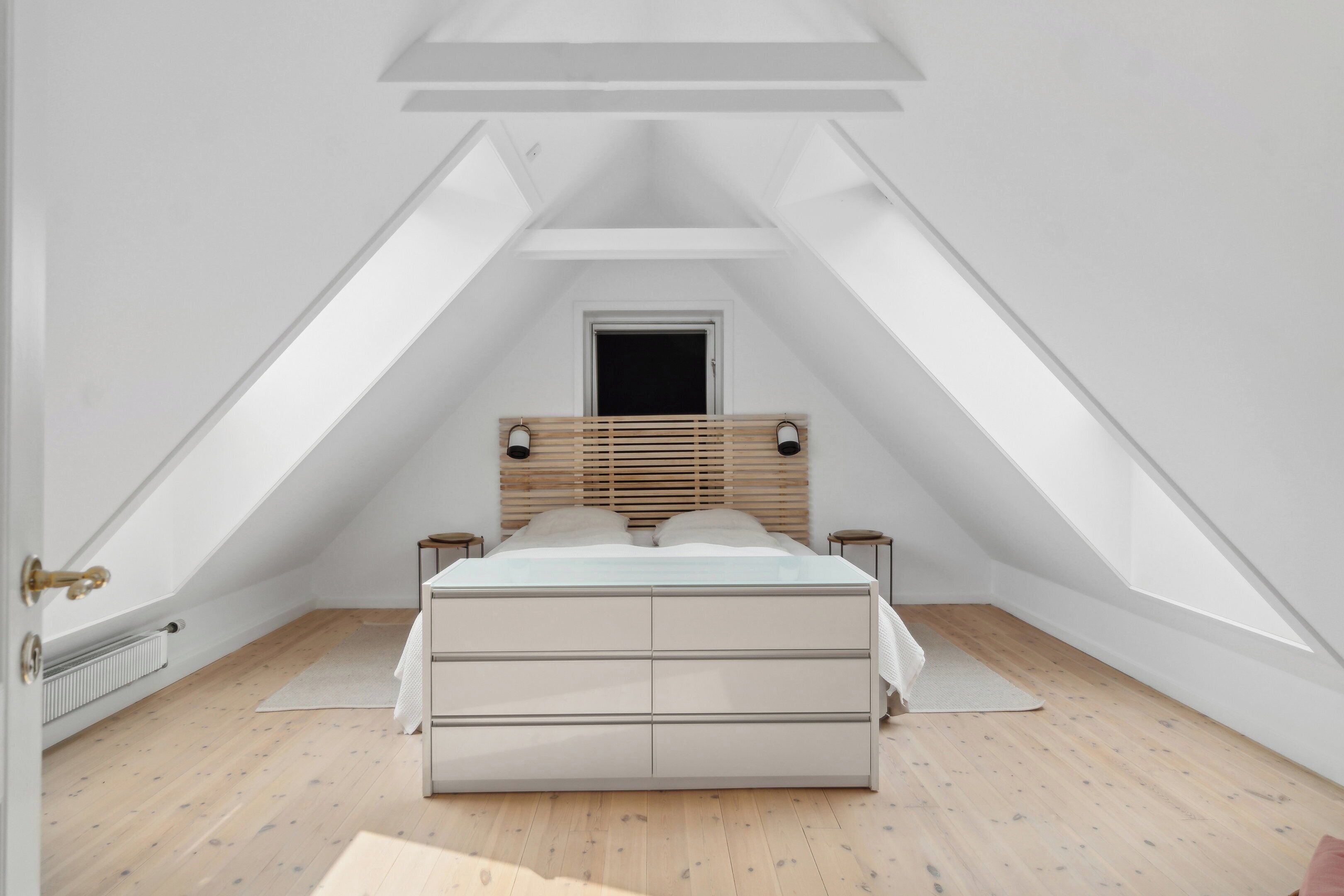 Une chambre avec un plafond en pente, un lit doté d’une tête de lit en bois, deux tables de chevet et un radiateur.