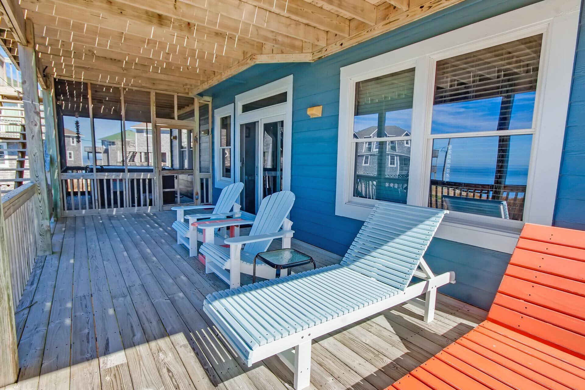 A covered outdoor deck with Adirondack chairs and a lounge chair, overlooking a body of water.