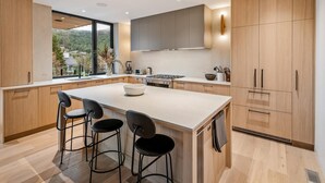 A modern kitchen with wooden cabinets, a central island with bar stools, a large window, and a view of the outdoors. - Blueberry Hill Retreat | Luxury, Close to everything, hot tub, incredible views, (Whistler)