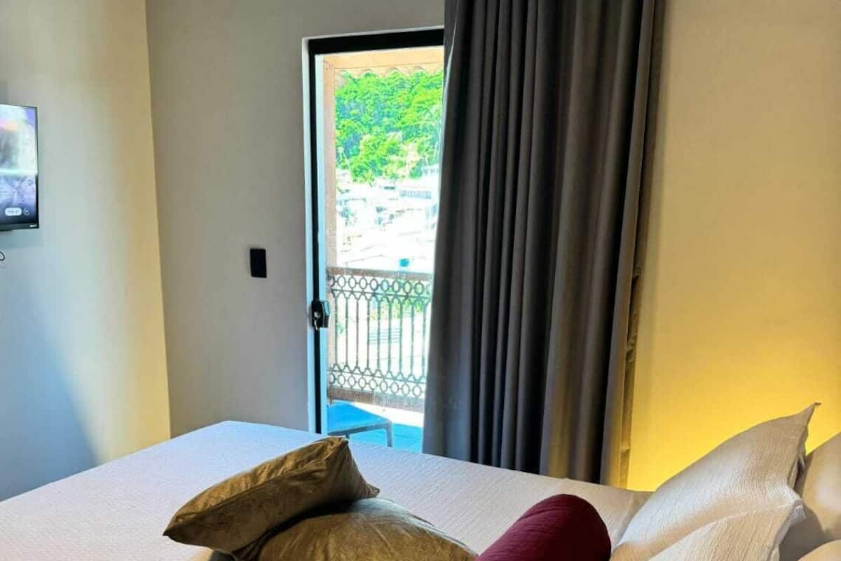 A neatly made bed with white linens and two pillows, a dark curtain partially drawn, and a view of a balcony with greenery outside.