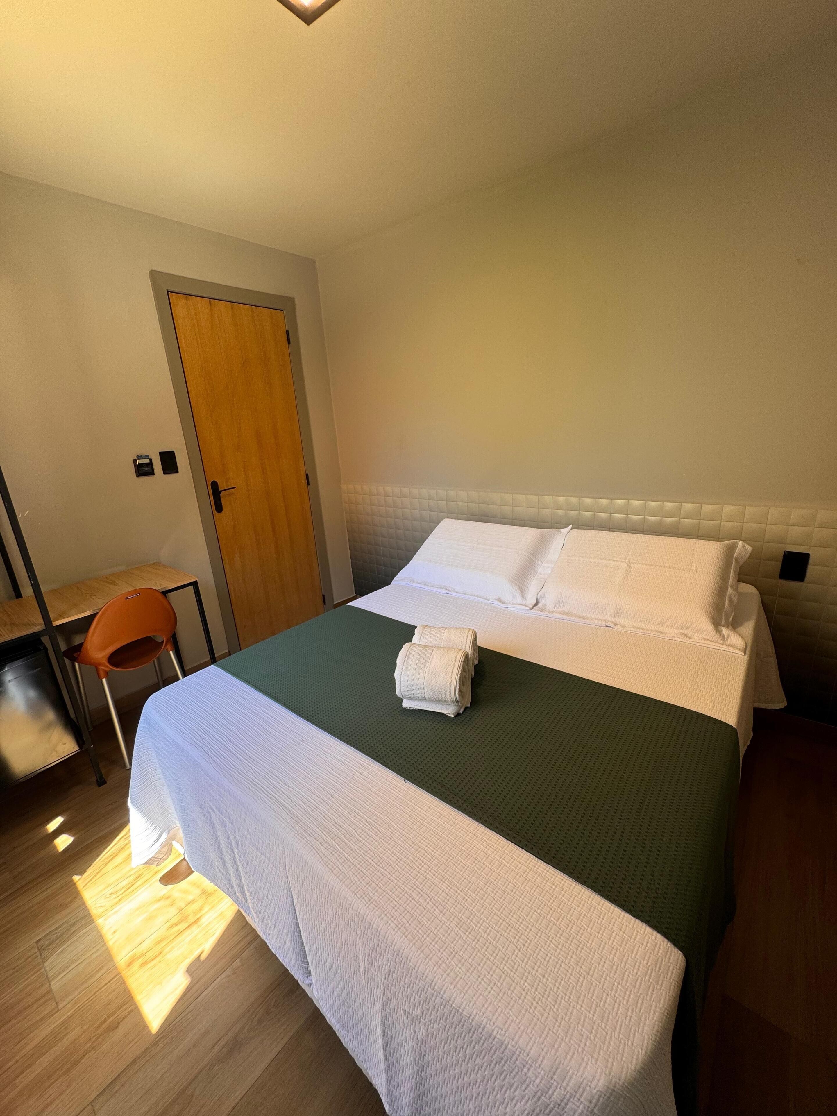 A neatly made bed with white linens, a green bedspread, and a rolled towel on the bed. There is a wooden desk with a chair, a door, and a small black object on the wall.