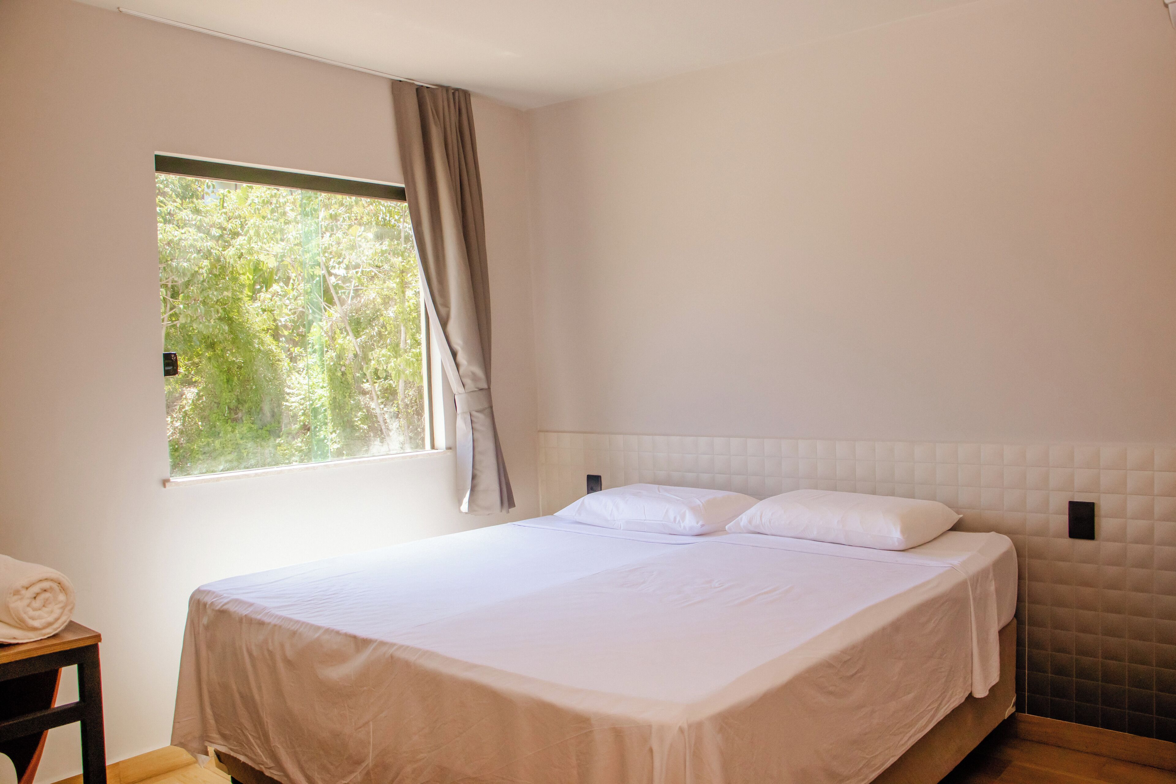 Una cama bien hecha en una habitación con ventana, una mesita de noche de madera y una lámpara fijada a la pared.