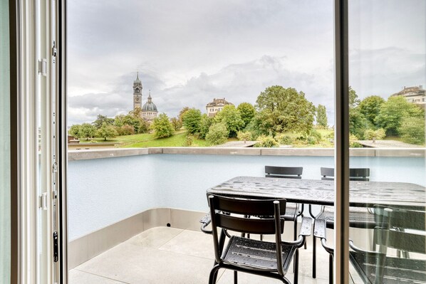 A balcony with a table and chairs overlooking a body of water, a park, and a historic building with a clock tower.