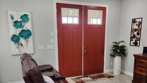 Una sala de estar con un sofá de cuero marrón, una puerta roja, un ventilador de techo y un cuadro con flores.