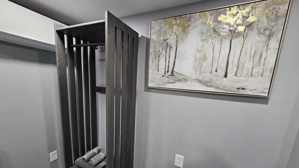 Ein Zimmer mit grauer Wand, einem Waldgemälde, einer hölzernen Bank mit Handtüchern und einem Mülleimer.