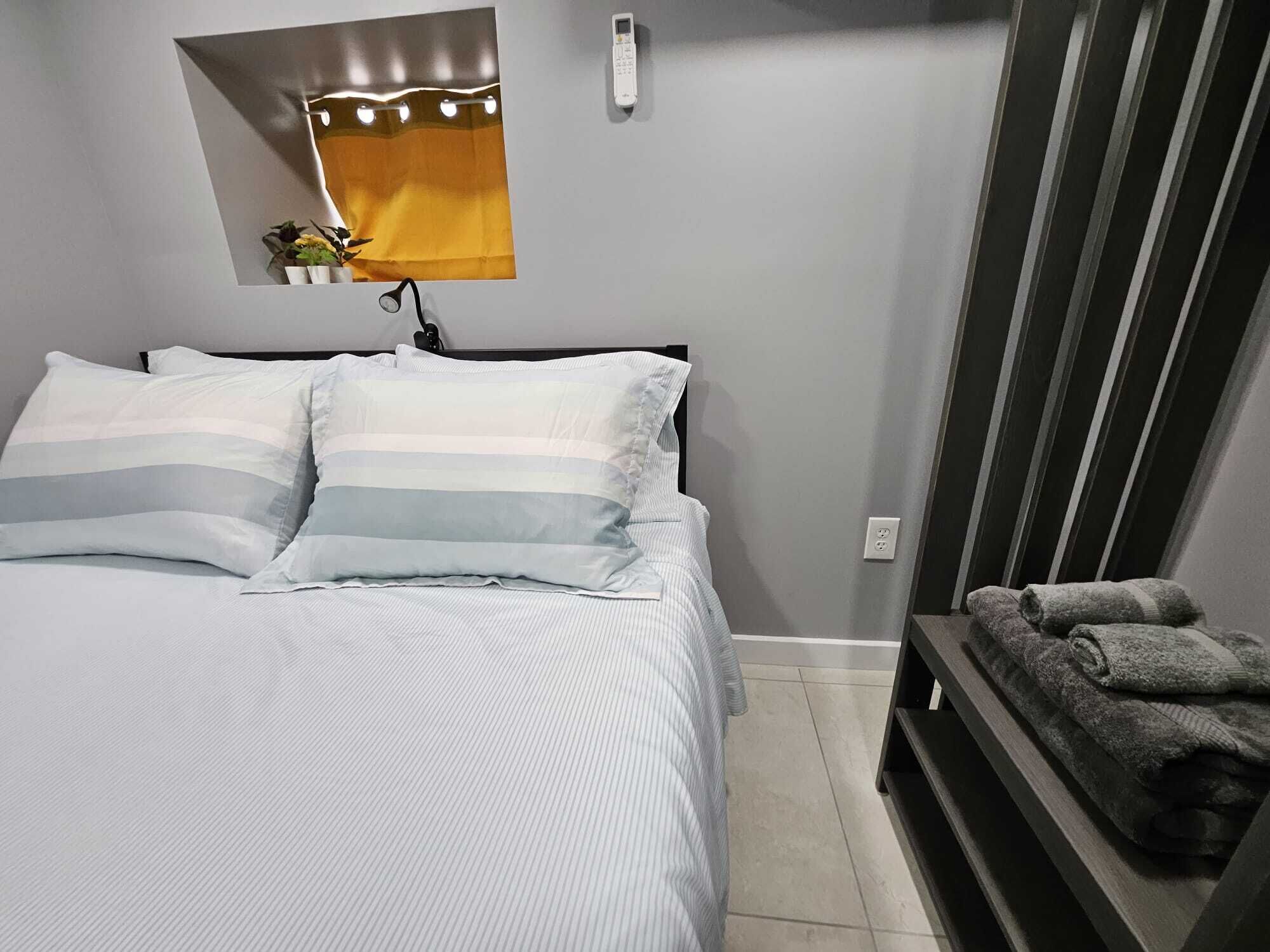 Cama bem arrumada com travesseiros listrados, um pequeno espelho iluminado, um telefone e um banco com toalhas.
