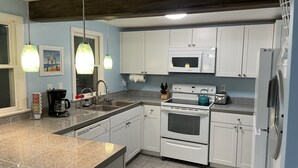 A modern kitchen with white cabinets, a stainless steel sink, a microwave, an oven, and a refrigerator. There are two pendant lights above the sink and a window with a view of the outside.