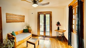 A cozy living room with a wooden sofa, a small table, a ceiling fan, and a large window with a view of greenery. - Apartamentos Casa Le Parc (Bocas del Toro)