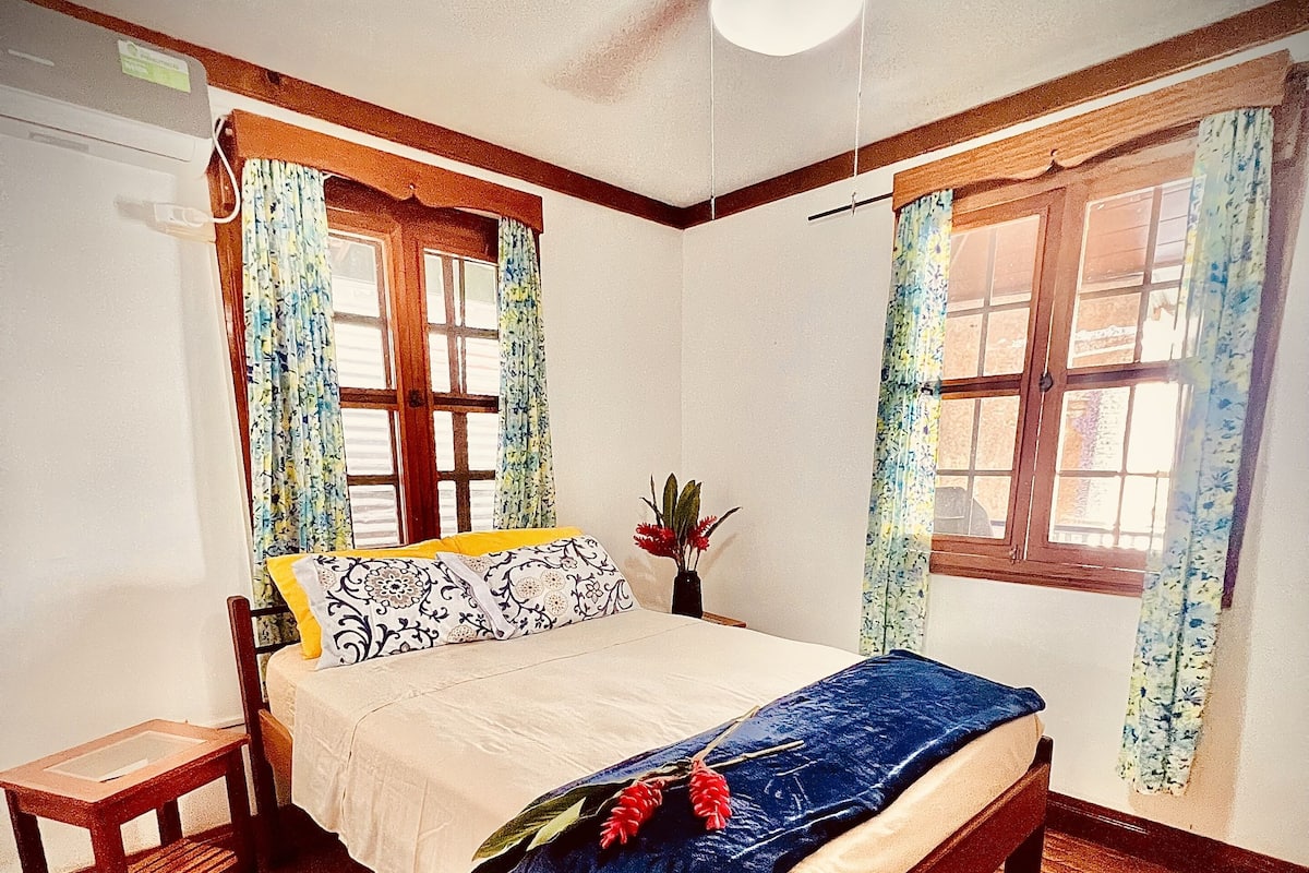Une chambre à coucher avec un lit en bois, une table de chevet, un ventilateur de plafond et deux fenêtres ornées de rideaux à motifs floraux.