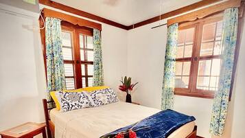 A bedroom with a wooden bed, a bedside table, a ceiling fan, and two windows with floral curtains.