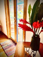 Una mesa de madera con un jarrón de flores rojas, un tapete tejido colorido y vistas a un balcón con barandilla.