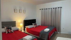 Une chambre d’hôtel avec deux lits, chacun recouvert d’une courtepointe rouge et d’une couverture grise. Deux tableaux encadrés ornent le mur, une lampe se trouve sur la table de chevet et une fenêtre est agrémentée de rideaux.