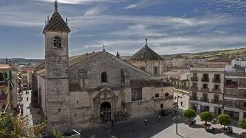 Una iglesia de piedra histórica con un campanario y una cúpula central, rodeada por una plaza con árboles y algunas personas.
