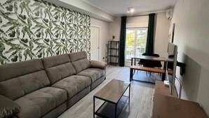 Una sala de estar con un sofá, una mesa de centro y un comedor con una mesa y sillas. Hay una ventana con cortinas, una planta y un televisor sobre un mueble.