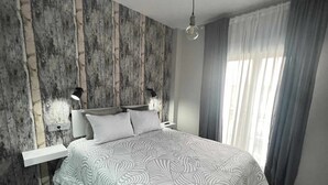 Un dormitorio moderno con un panel de pared de madera, una cama grande con ropa de cama estampada, lámparas de noche y una ventana con cortinas.