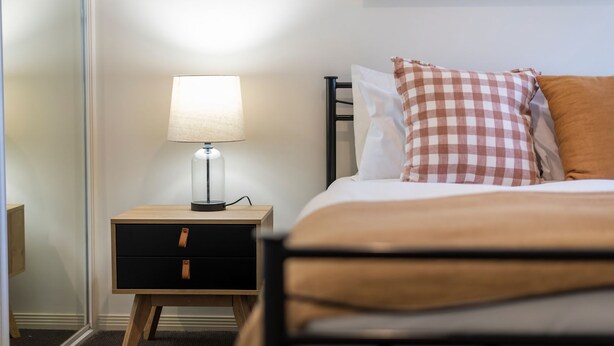 A neatly made bed with a checkered pillow and a bedside lamp on a wooden nightstand with drawers. - Breakers 1/8- Stone Real Estate Yamba (Yamba)