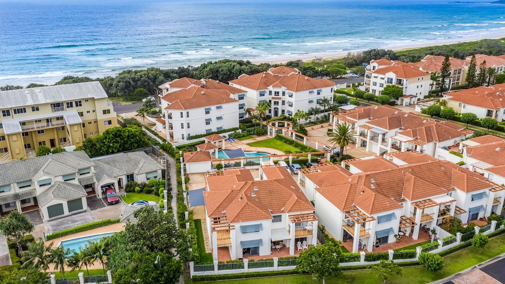 A coastal residential area with multiple two-story buildings, a swimming pool, and a beach in the background.