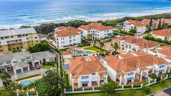 A coastal residential area with multiple two-story buildings, a swimming pool, and a beach in the background.