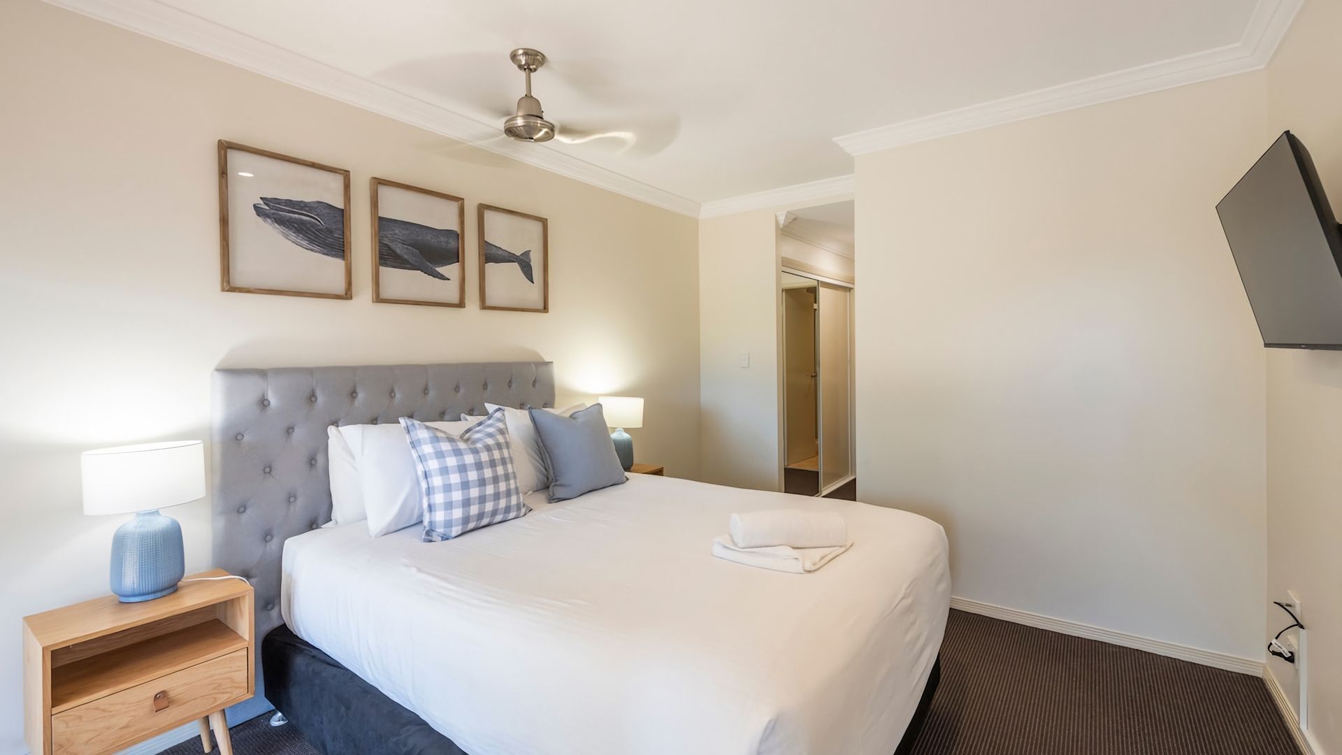 Une chambre d’hôtel avec un grand lit, une table de chevet, une lampe et un ventilateur de plafond. Trois tableaux encadrés sont accrochés au mur au-dessus du lit.