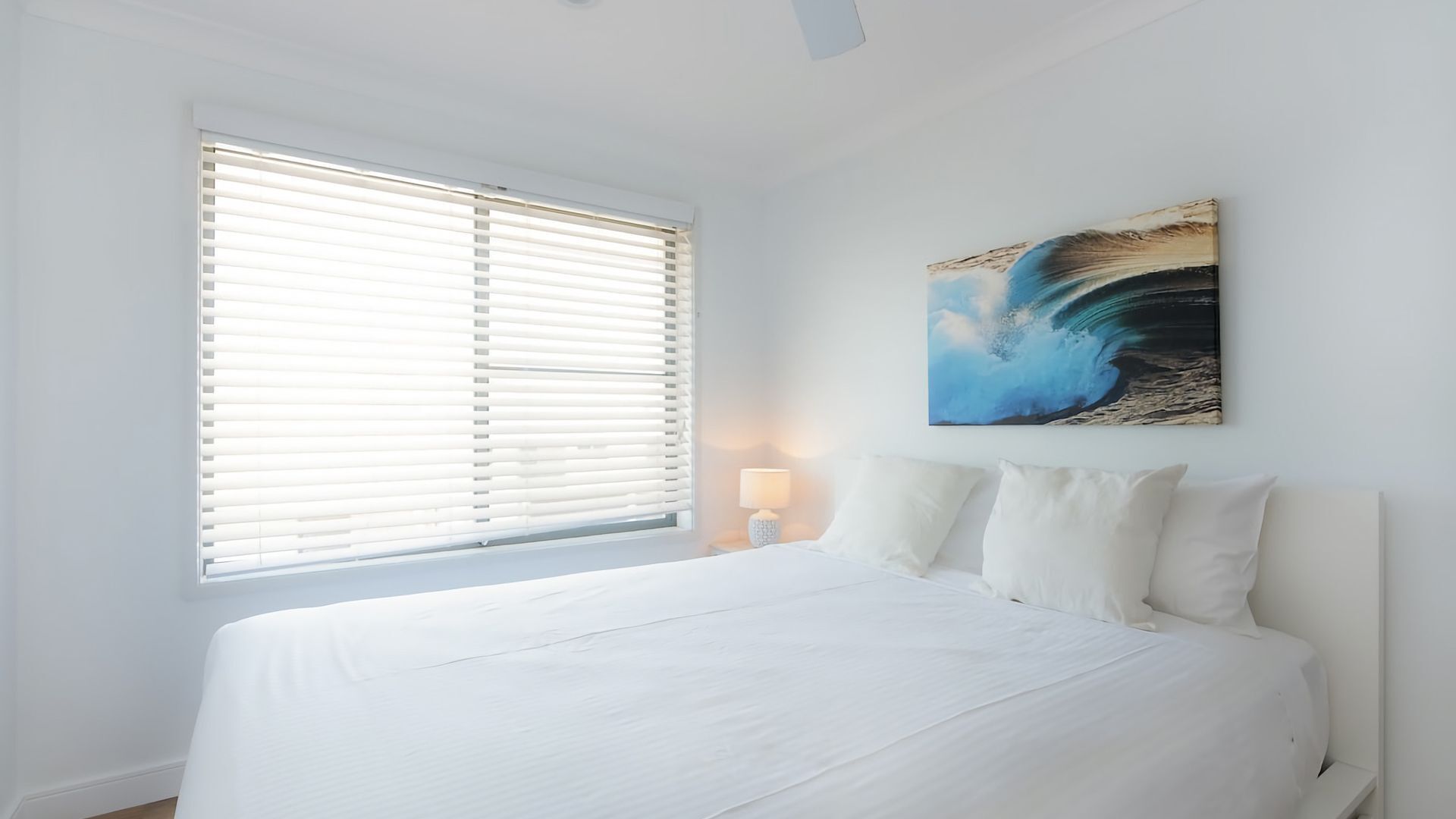 Een slaapkamer met een groot bed, een raam met zonwering, een bedlampje en een aan de muur hangend schilderij.