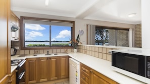 A kitchen with wooden cabinets, a built-in oven, a dishwasher, and a large window offering a view of a lake. - Peninsula Court 6- Stone Real Estate Yamba (Yamba)