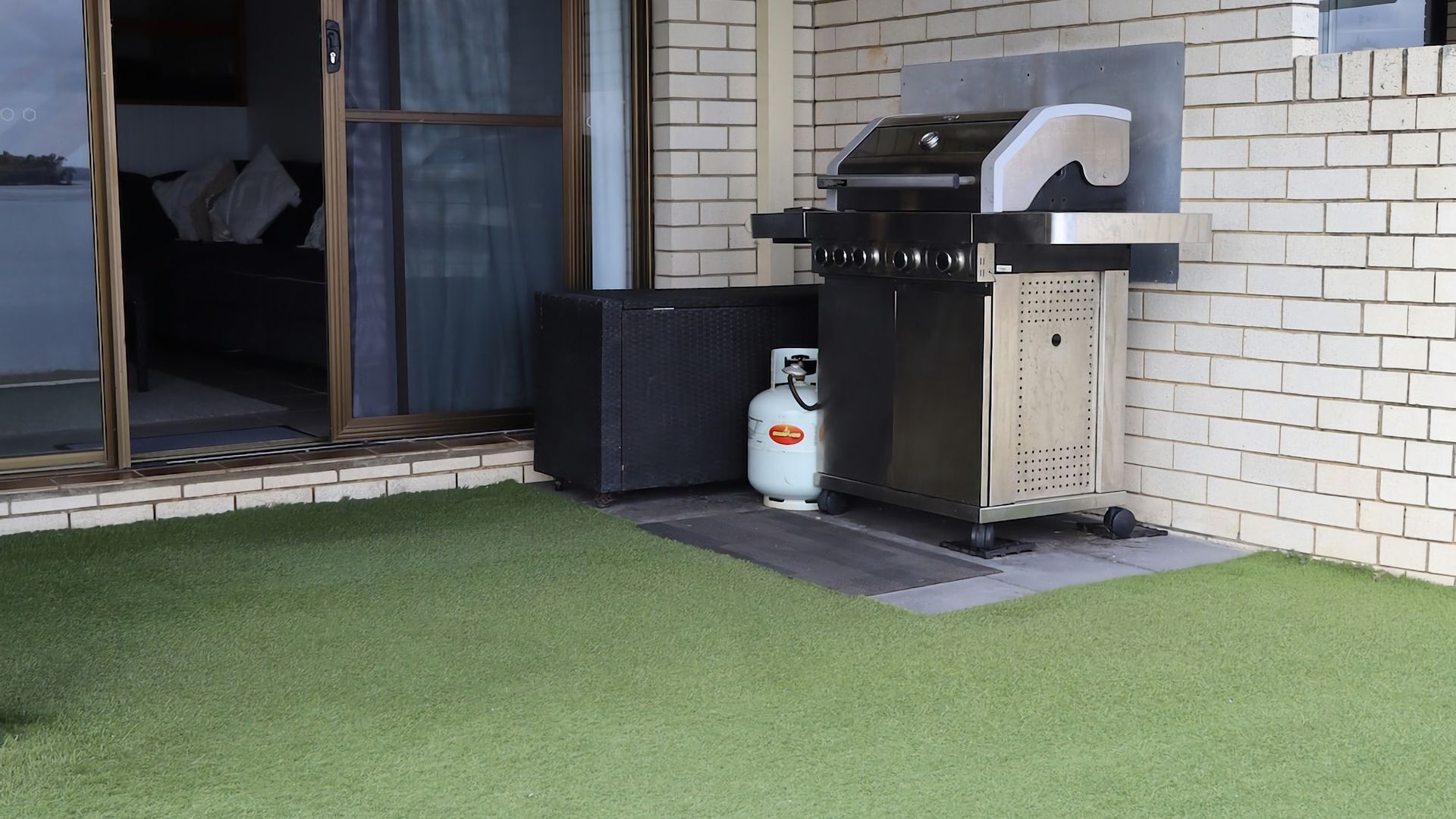 An outdoor grill area with a gas cylinder, a trash bin, and a sliding glass door leading to a room.