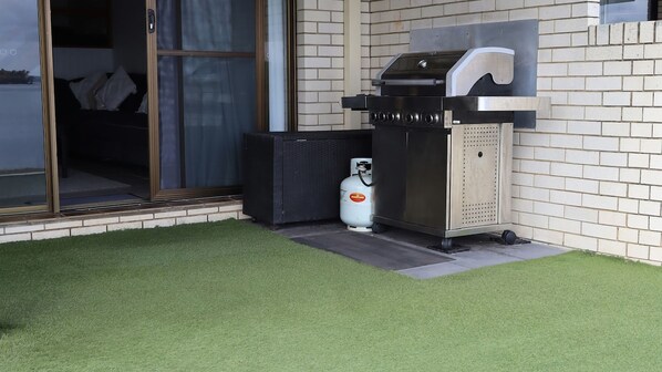 An outdoor grill area with a gas cylinder, a trash bin, and a sliding glass door leading to a room.
