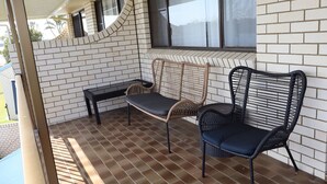 Un espacio de estar al aire libre cubierto, con dos sillones de mimbre y un banco, un muro de ladrillo y una ventana.