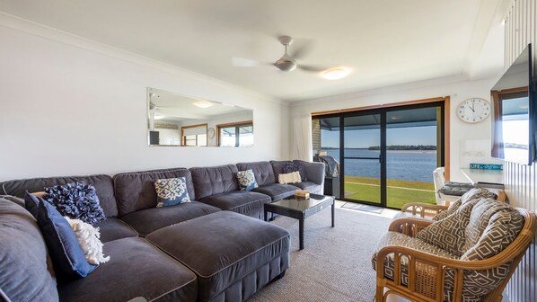 A living room with a large sectional sofa, a coffee table, and a wicker chair. There is a sliding glass door leading to a balcony with a view of the water.