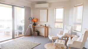 A modern living room with a glass door leading to an outdoor area, a tripod, a bookshelf, and a seating area with ottomans and a table. - Breakers 2/12- Stone Real Estate Yamba (Yamba)