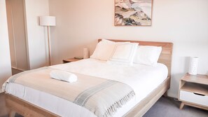 A neatly made bed with white linens and a rolled towel, a bedside table with a lamp, and a wall-mounted painting.