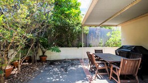 Une terrasse couverte avec une table et des chaises, entourée de plantes en pot et d’une clôture en bois.