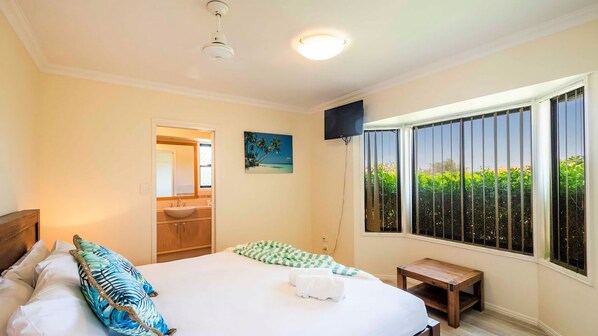 Une chambre à coucher comprenant un lit, une table de chevet, une fenêtre donnant sur un espace verdoyant, et une salle de bain avec un lavabo et un miroir.