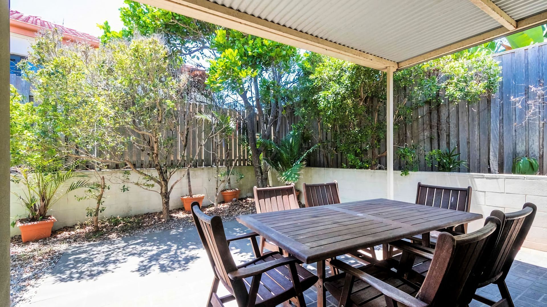 A covered patio with a wooden table and chairs, surrounded by potted plants and trees.