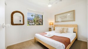 A hotel room with a bed, a bedside lamp, a framed picture, a mirror, and a window with a view of greenery and a building. - Kailani Beach House - Stone Real Estate Yamba (Yamba)