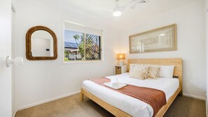 A hotel room with a bed, a bedside lamp, a framed picture, a mirror, and a window with a view of greenery and a building. - Kailani Beach House - Stone Real Estate Yamba (Yamba)
