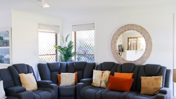 A modern living room with a sectional sofa, a decorative mirror, and a view through a window with a plant. - Kailani Beach House - Stone Real Estate Yamba (Yamba)