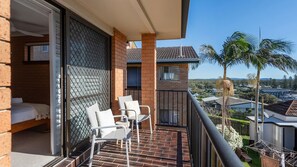 A balcony with a bed, two chairs, and a view of a residential area with houses and palm trees. - Elsewhere 3- Stone Real Estate Yamba (Yamba)