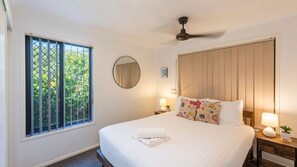 A hotel room with a large bed, a ceiling fan, a window with a view of greenery, and a bedside table with a lamp and a plant. - 32 Beachside Way -Stone Real Estate Yamba (Yamba)