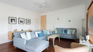 Un salon avec un canapé d’angle, une petite table et un fauteuil beige. Les murs sont ornés de tableaux encadrés, il y a un ventilateur de plafond et une fenêtre avec des stores.