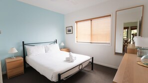 A bedroom with a bed, bedside tables, a window with blinds, a mirror, and a lamp. - Six - River Stone Real Estate Yamba (Yamba)