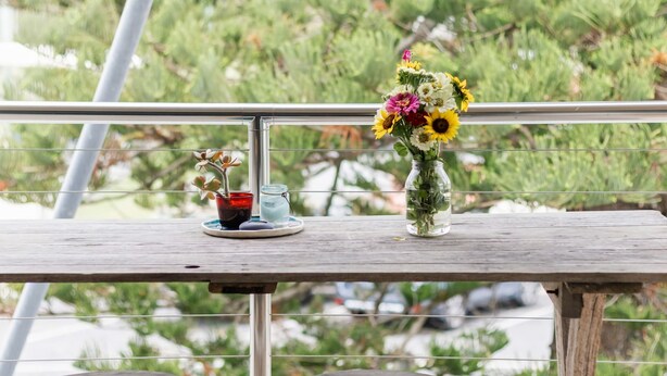 Uma mesa de madeira com um vaso de vidro com flores e um copo de água numa varanda.