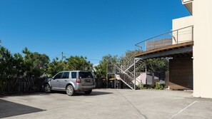 Un espace de stationnement avec une voiture, un escalier menant à un bâtiment et une clôture bordée d’arbres.